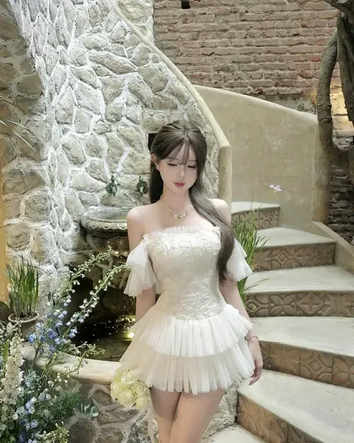 Nàng tóc ngắn mặc váy tầng màu kem đứng trên bậc đá, cúi nhìn nhẹ nhàng