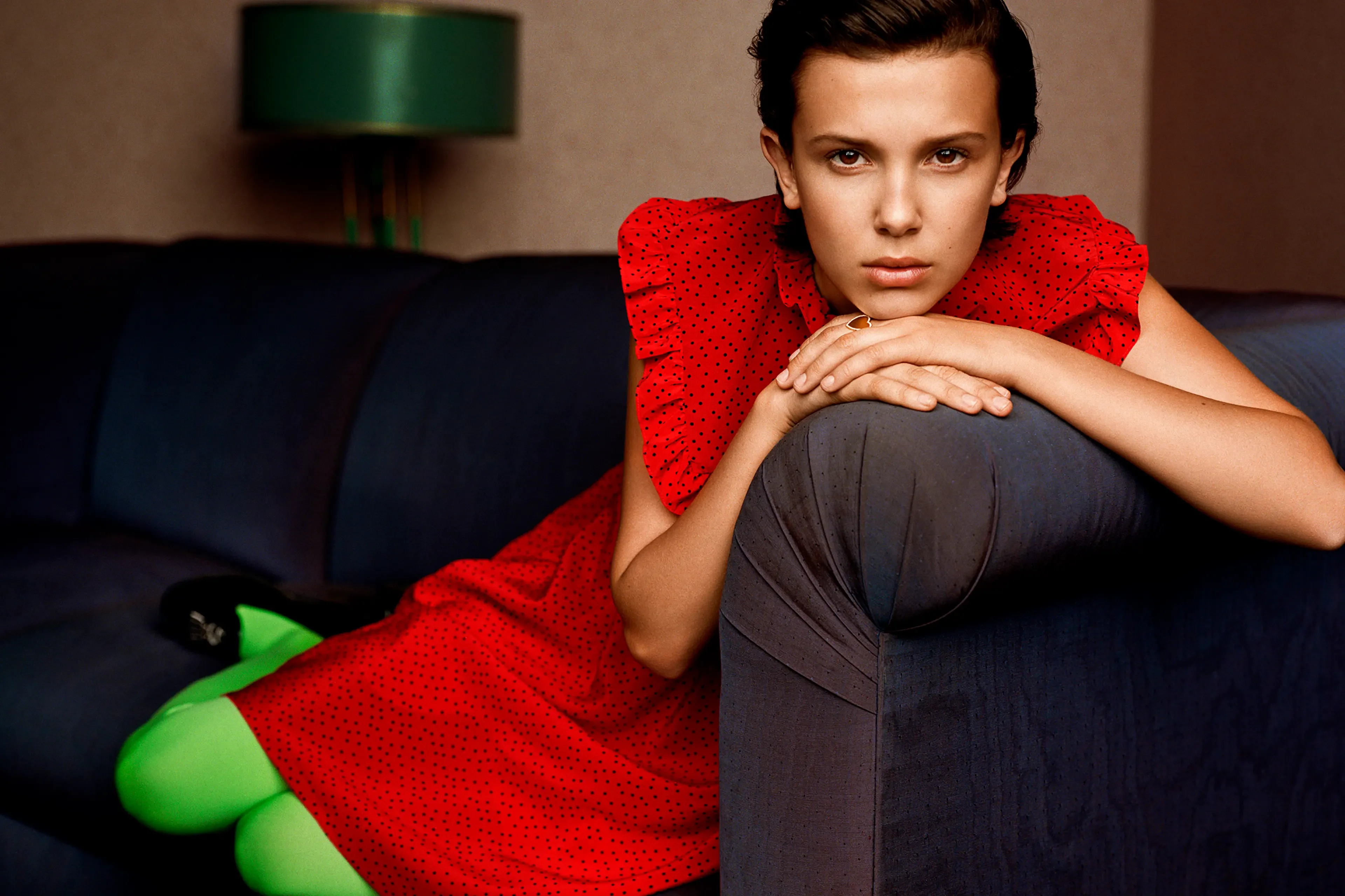 Ảnh chân dung thời trang Millie Bobby Brown mặc váy đỏ chấm bi và quần tất xanh neon trên ghế sofa